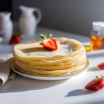 Pfannkuchen Rezept – Stapel dünner Pfannkuchen garniert mit Erdbeere und Puderzucker, serviert auf einem weißen Teller.